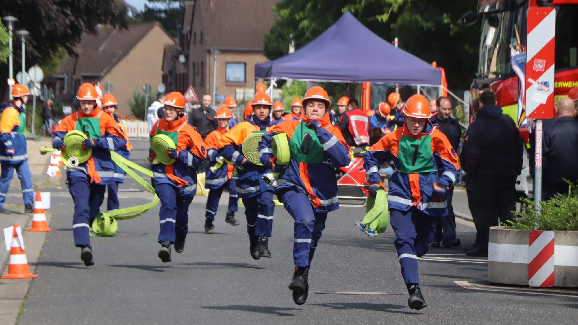 Auf dem Foto ist zu sehen, wie junge Feuerwehrleute mit Schläuchen zum fiktiven Einsatzort laufen.