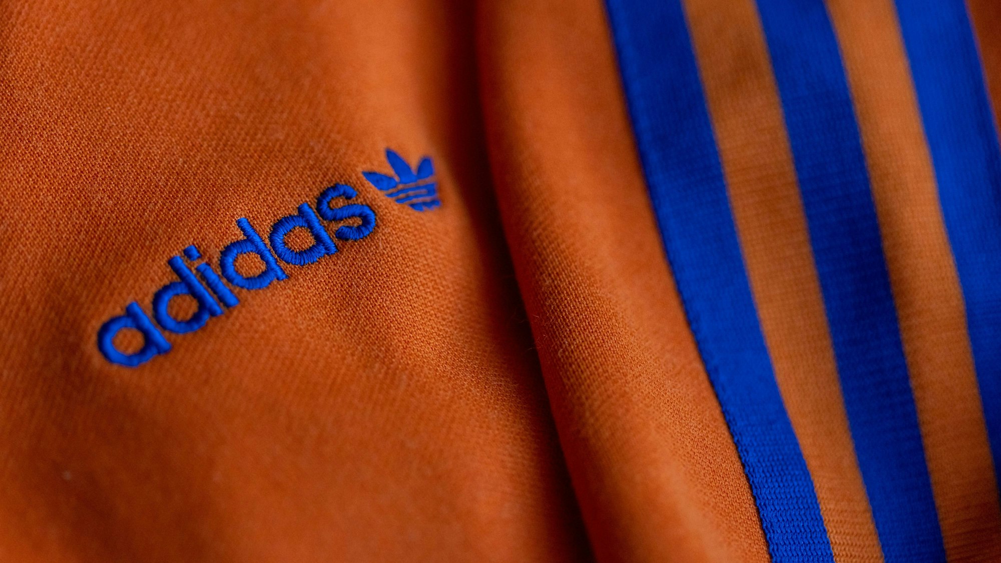 ARCHIV - 08.03.2022, Bayern, Herzogenaurach: Das Logo des Sportartikelherstellers adidas in blau auf einer orangenen Jacke. (zu dpa: «Urteil: Streifen auf Nike-Sporthose verletzen Adidas-Markenrechte») Foto: Daniel Karmann/dpa +++ dpa-Bildfunk +++