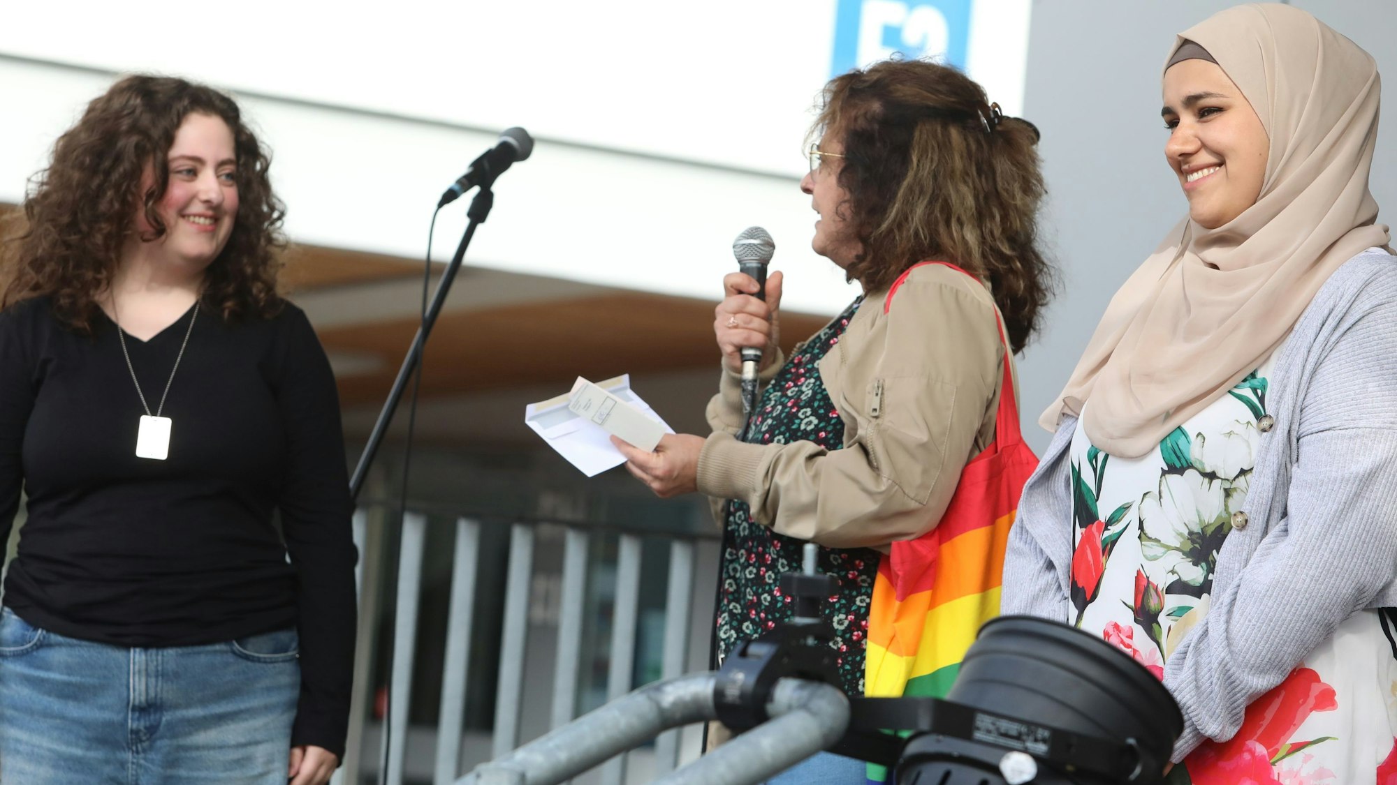 Drei Frauen stehen auf einer Bühne, eine hält ein Mikrofon in der Hand.