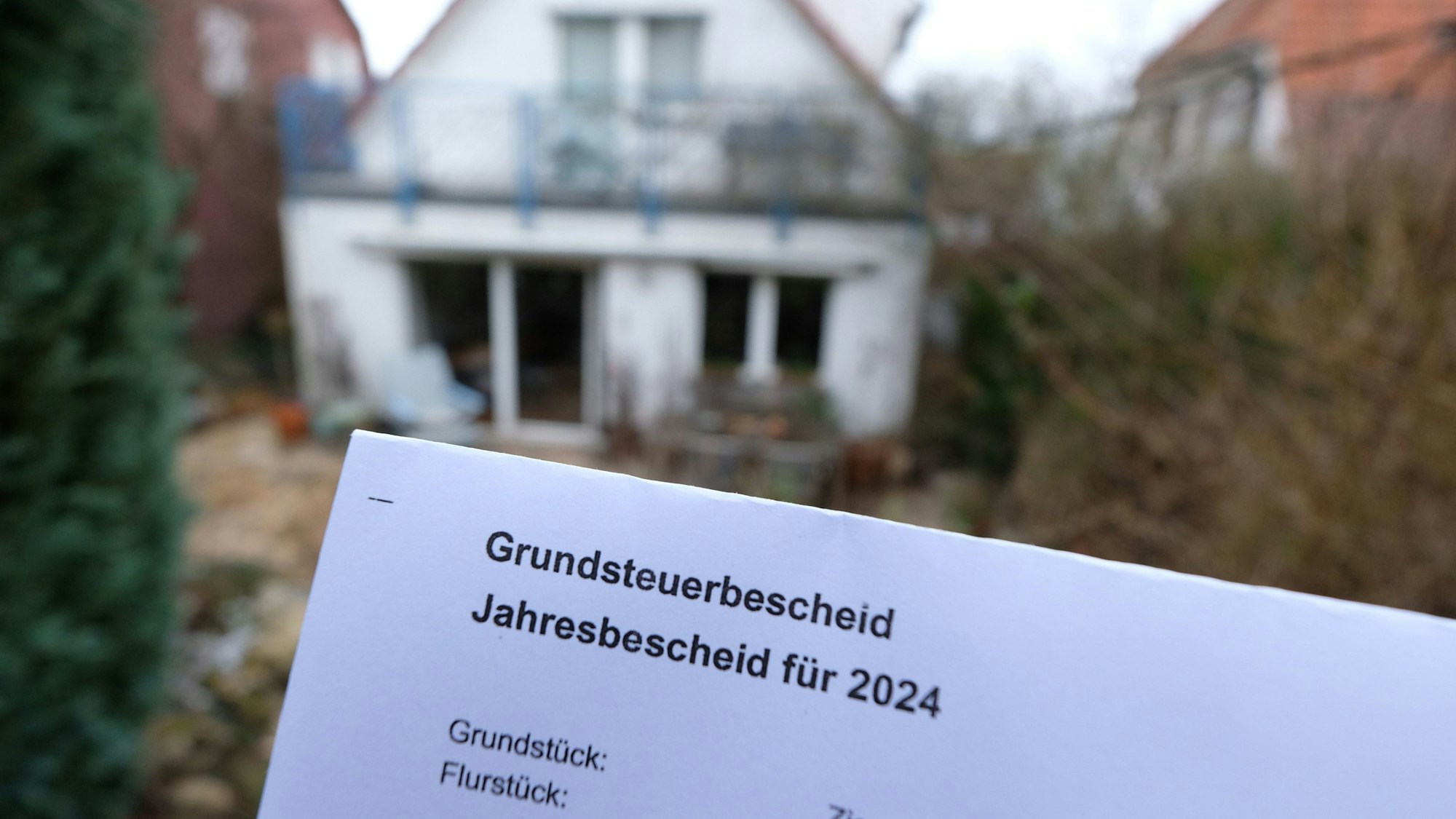 Ein Grundsteuerbescheid für 2024 wird vor einem Privatgrundstück hochgehalten.