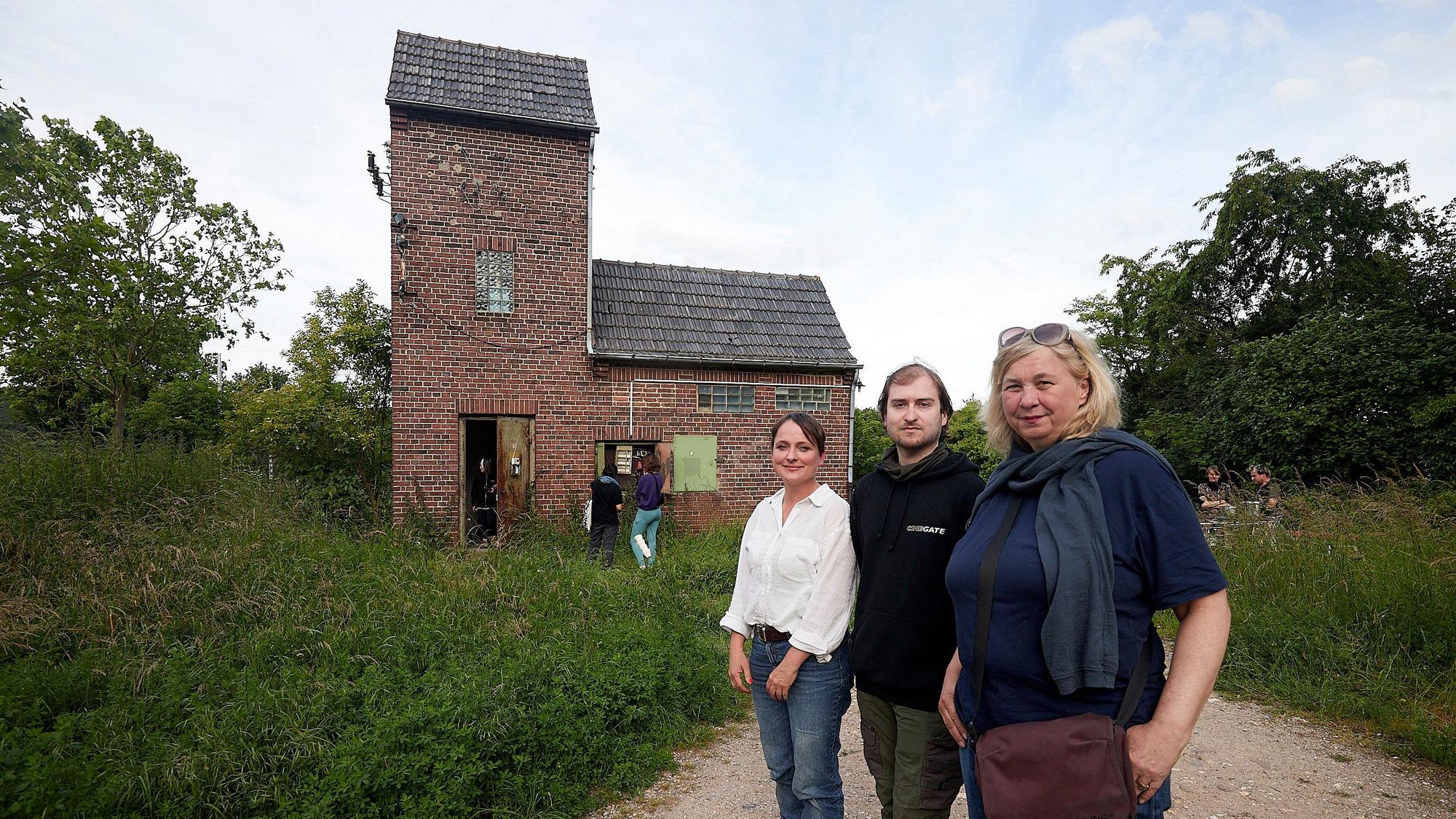 Zwei Frauen und ein Mann, Stefanie Klingemann, Pascal Dreier und Ute Hörner, stehen vor dem alten Trafohaus in Satzvey, das von vielen grünen Sträuchern und Bäumen umgeben ist.
