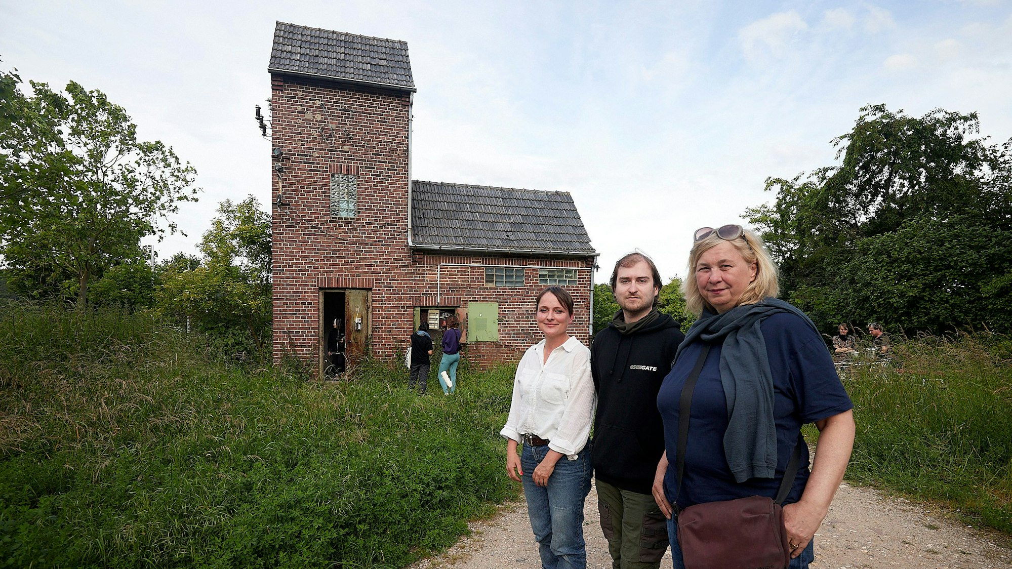 Zwei Frauen und ein Mann, Stefanie Klingemann, Pascal Dreier und Ute Hörner, stehen vor dem alten Trafohaus in Satzvey, das von vielen grünen Sträuchern und Bäumen umgeben ist.