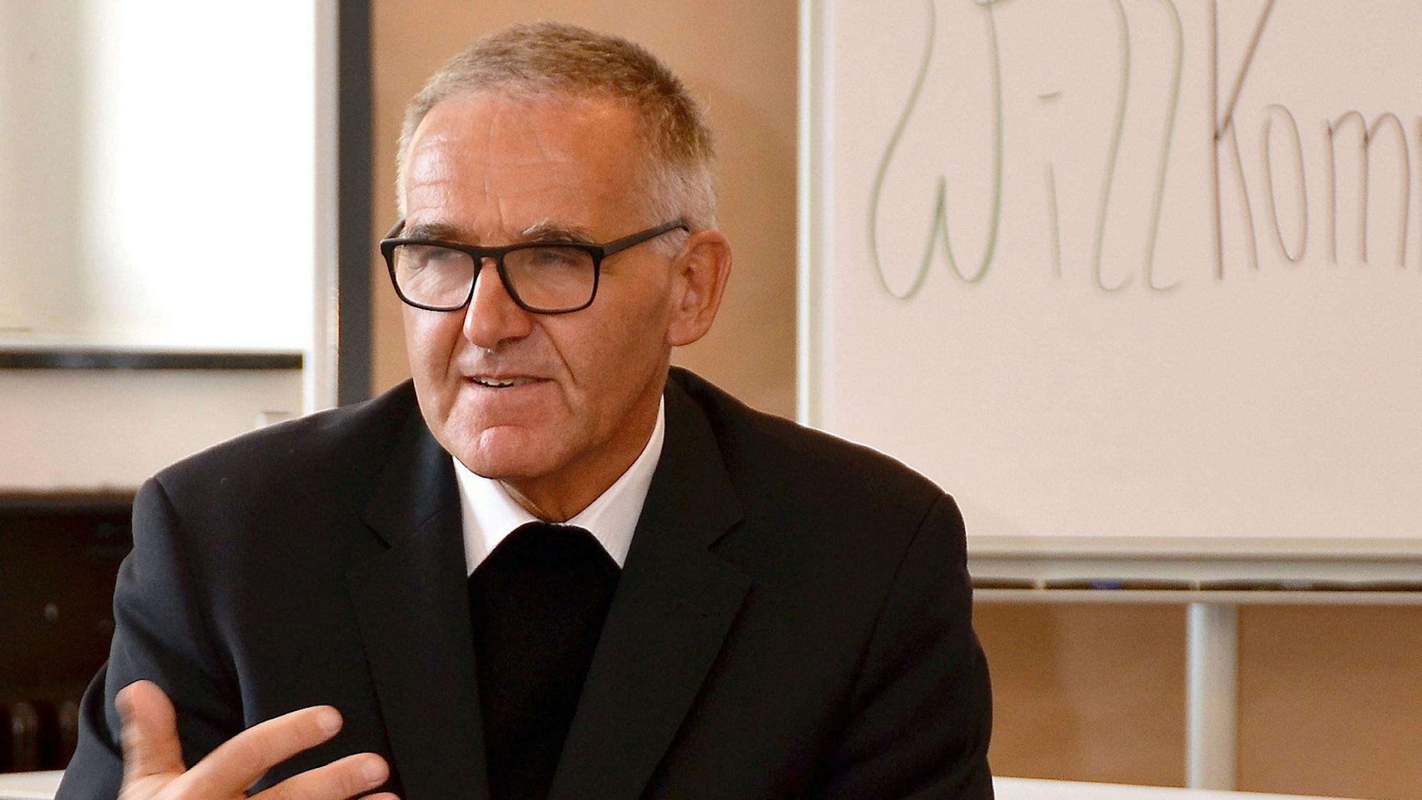 Dr. Johannes Bündgens sitzt vor einer Tafel, auf der die ersten Buchstaben des Wortes „Willkommen“ zu lesen sind.