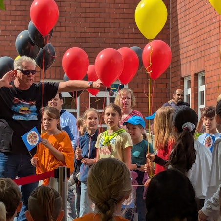Das Foto zeigt einen Sänger in Pose vor Schulkindern, die Luftballons in der Hand halten.