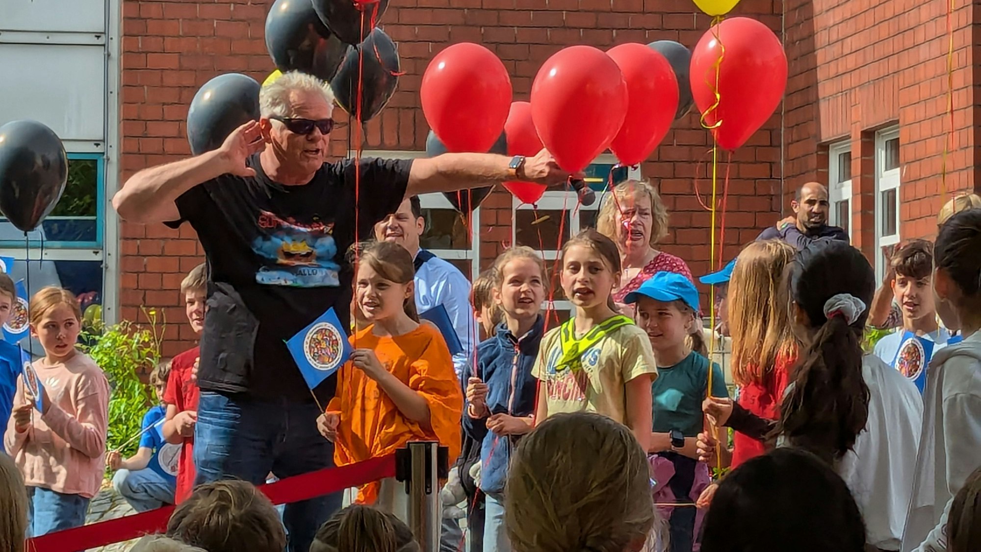 Das Foto zeigt einen Sänger in Pose vor Schulkindern, die Luftballons in der Hand halten.