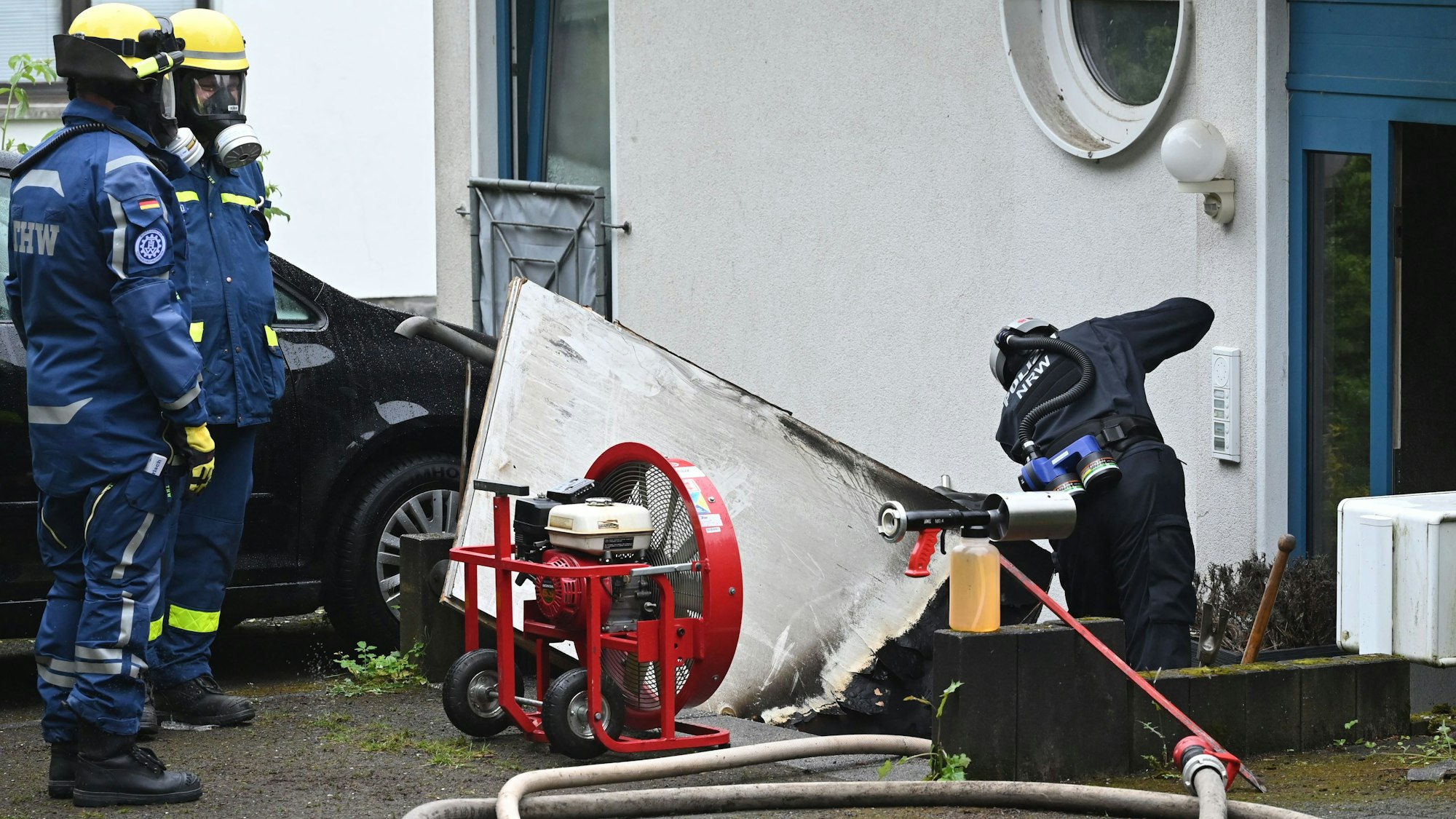 Ein Polizist steht vor der Eingangstür zu einem Mehrfamilienhaus. Zwei THW-Kräfte schauen ihm zu.