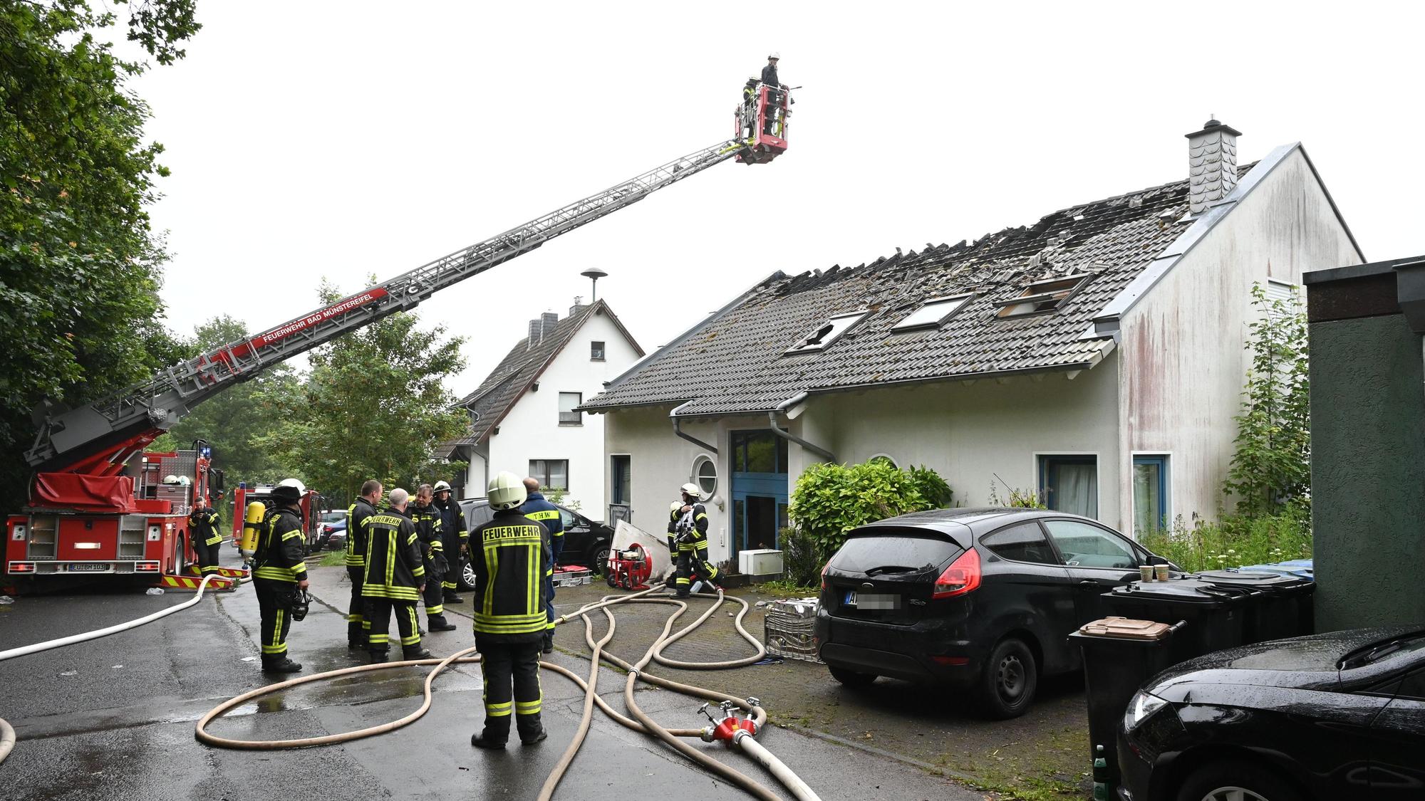 Zahlreiche Feuerwehrleute stehen vor einem Haus in Bad Münstereifel-Holzem. Eine Drehleiter ist ausgefahren, das Dach massiv beschädigt. Das Bild stammt vom Juni 2024.