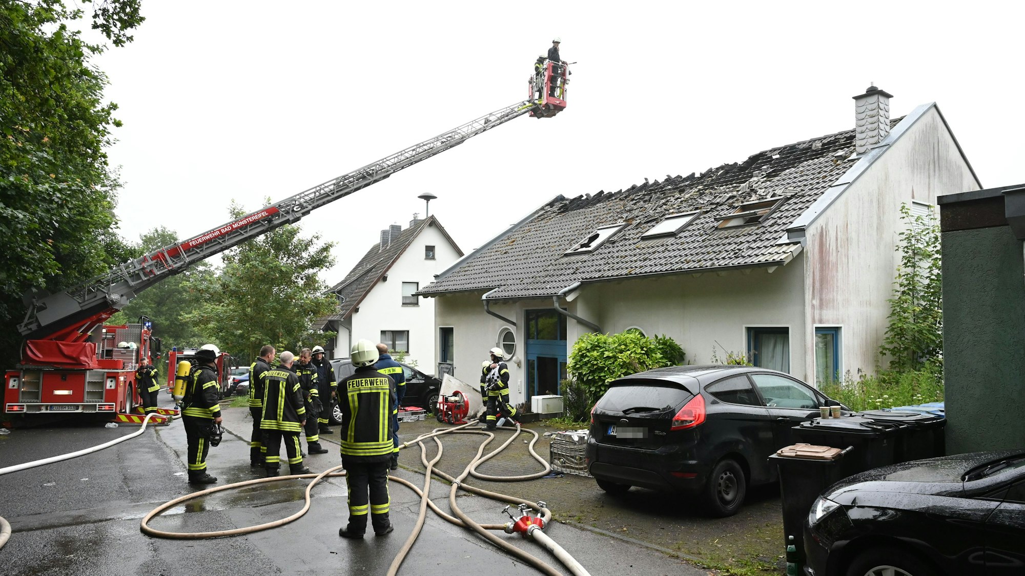Zahlreiche Feuerwehrleute stehen vor einem Haus in Bad Münstereifel-Holzem. Eine Drehleiter ist ausgefahren, das Dach massiv beschädigt. Das Bild stammt vom Juni 2024.