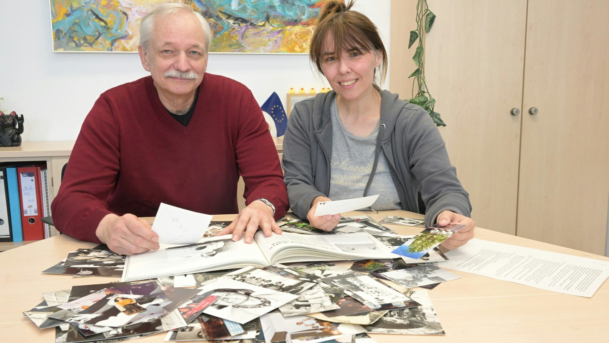 Müller und Matthes sitzen am Tisch, auf dem viele Fotos und die Schulchronik liegt.