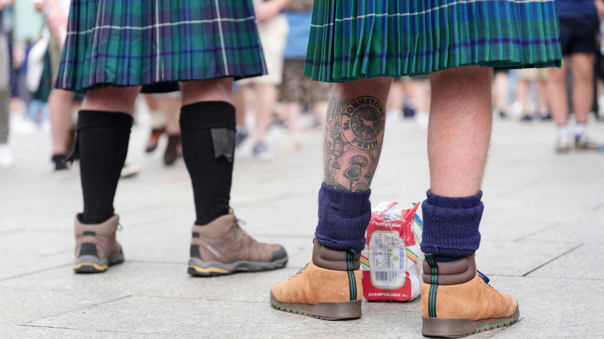 Zwei Männer mit blau-grünen Kilts und Wanderschuhen stehen in der Kölner Innenstadt, man sieht ihre Beine und Füße von hinten.