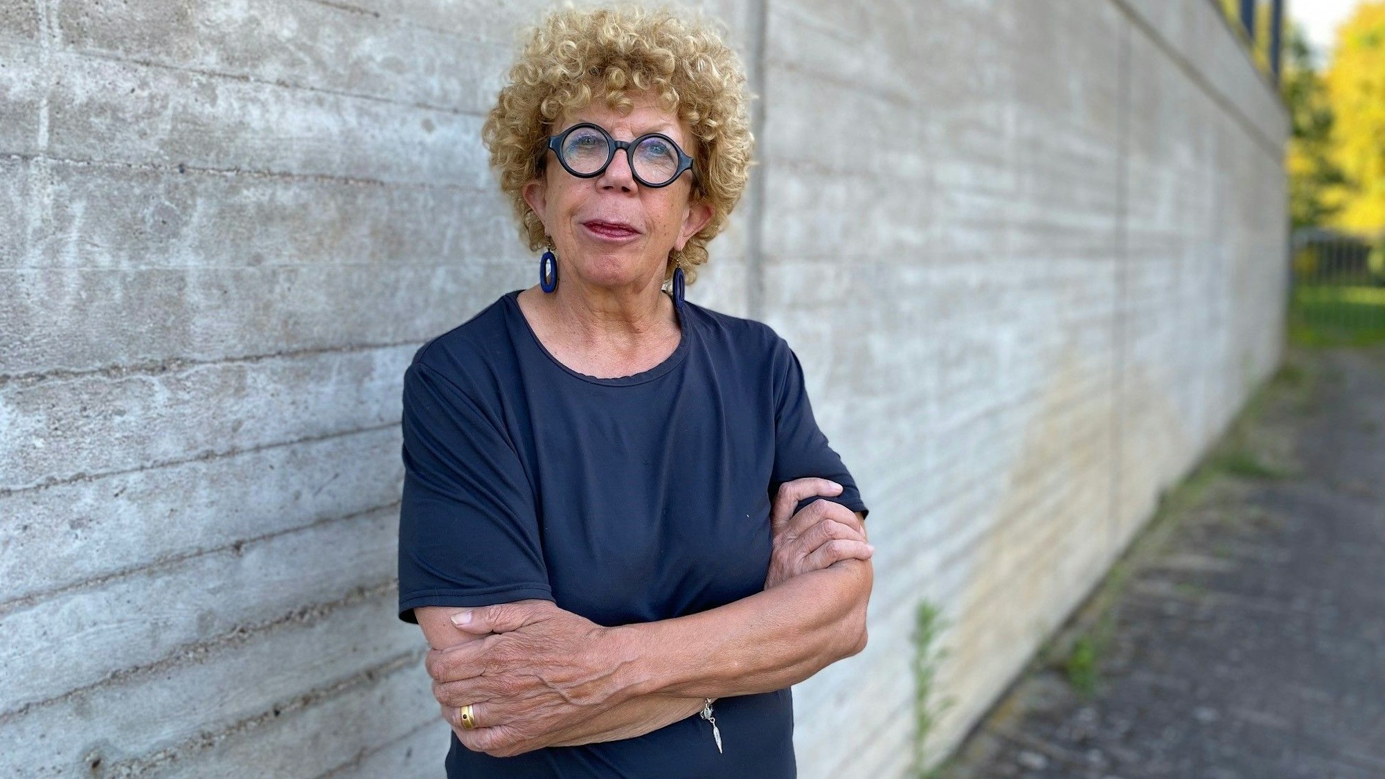 Auf dem Foto ist eine Frau zu sehen, die mit verschränkten Armen vor einer Mauer steht.