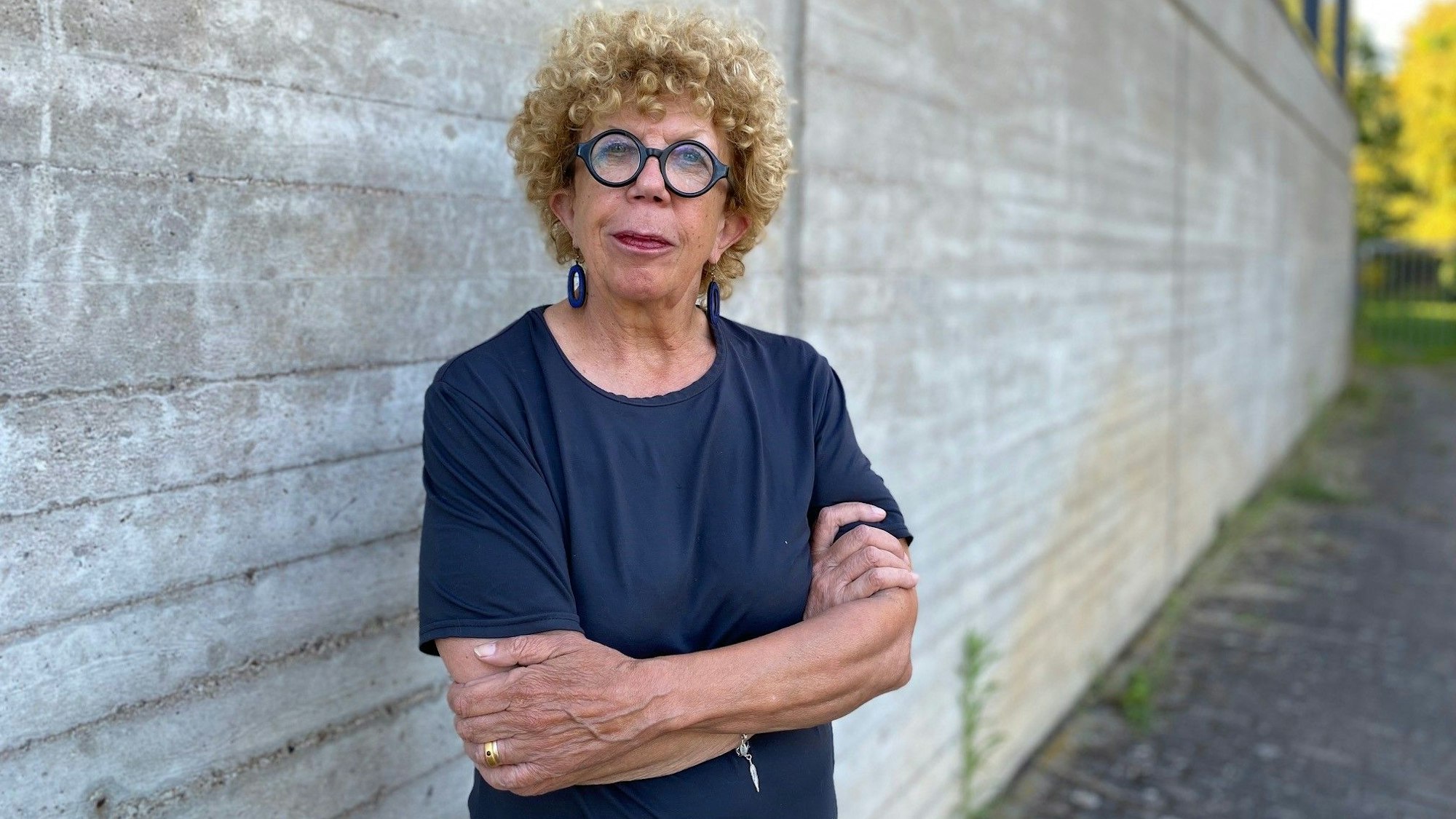 Auf dem Foto ist eine Frau zu sehen, die mit verschränkten Armen vor einer Mauer steht.