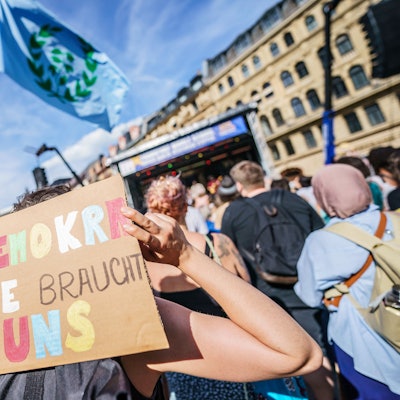 Ein Plakat mit der Aufschrift „Demokratie braucht uns“ wird hochgehalten.