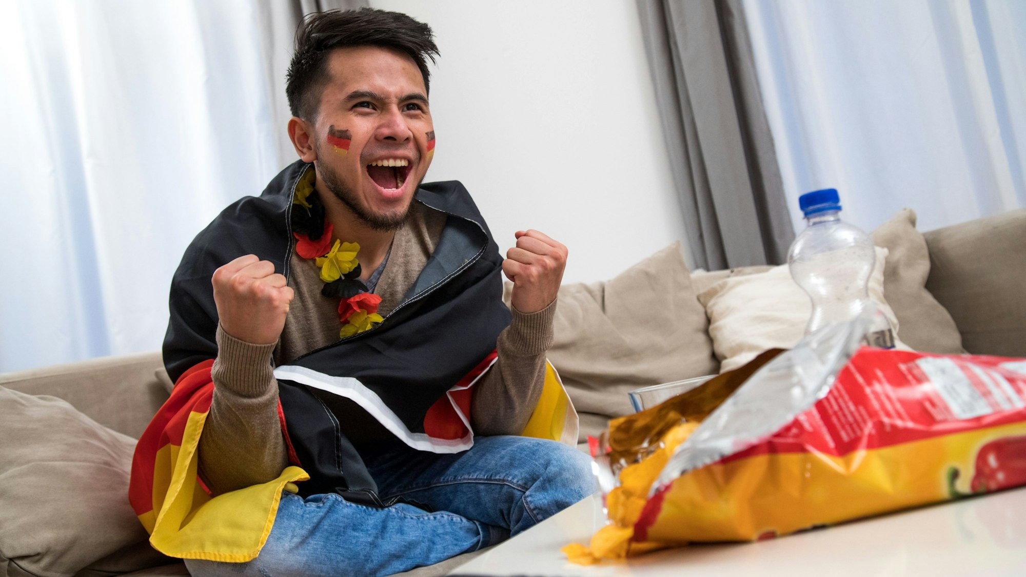 Ein Mann mit Deutschlandfahne sitzt auf dem Sofa und freut sich.