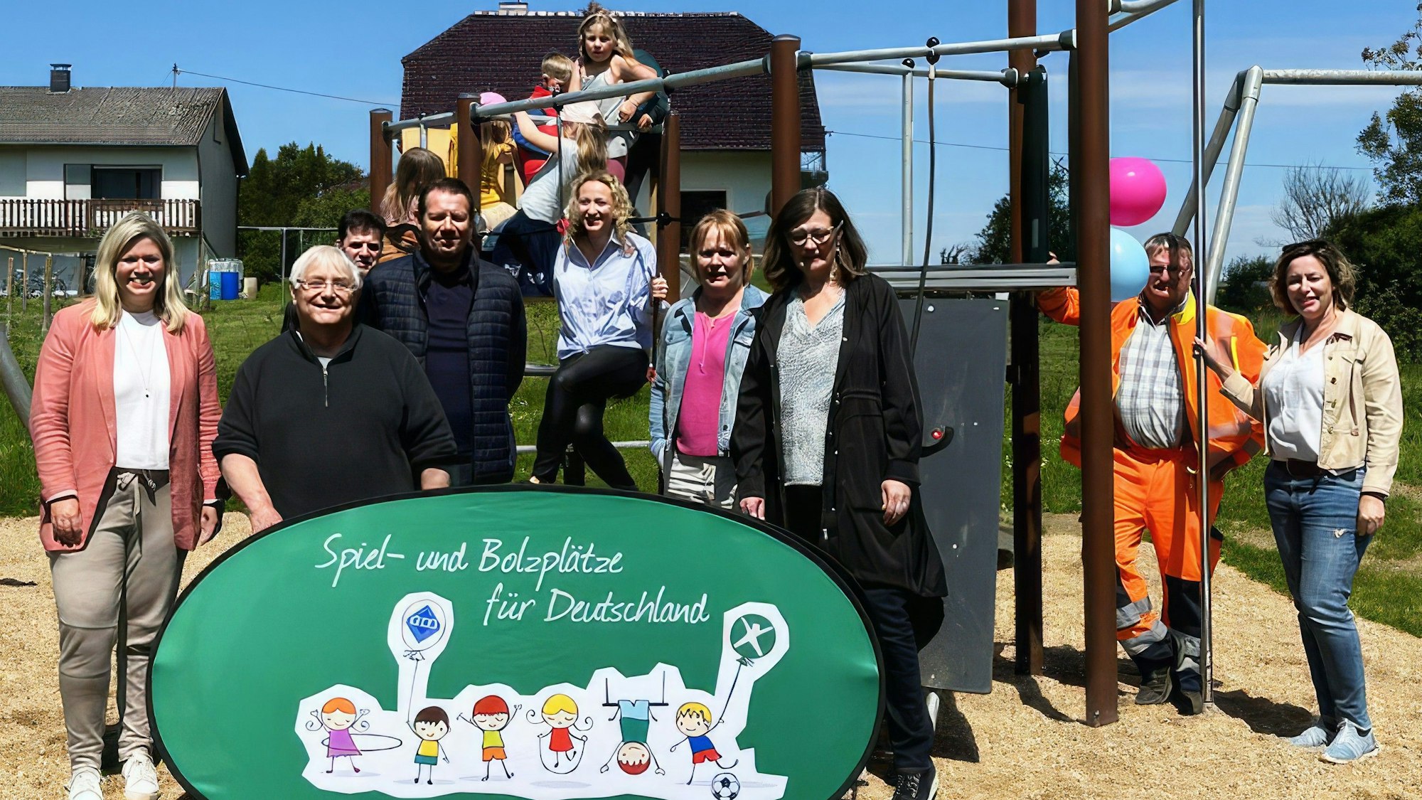 Erwachsene und Kinder posieren auf und um ein Klettergerüst auf einer Sandfläche. Vor ihnen steht ein Schild mit der Aufschrift: Spiel- und Bolzplätze für Deutschland.