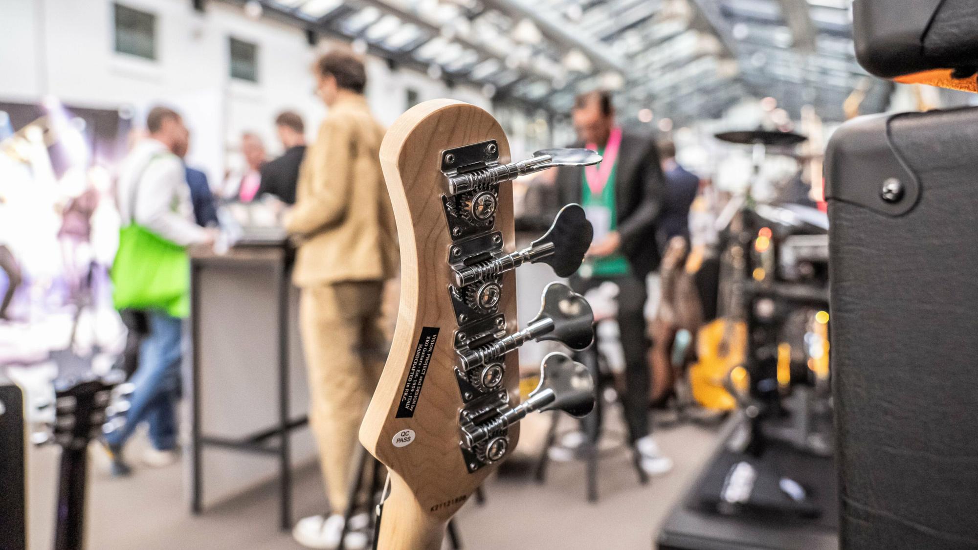 Teile von einer Gitarre sind im Vordergrund, im Hintergrund halten sich mehrere Personen in einer Halle auf.