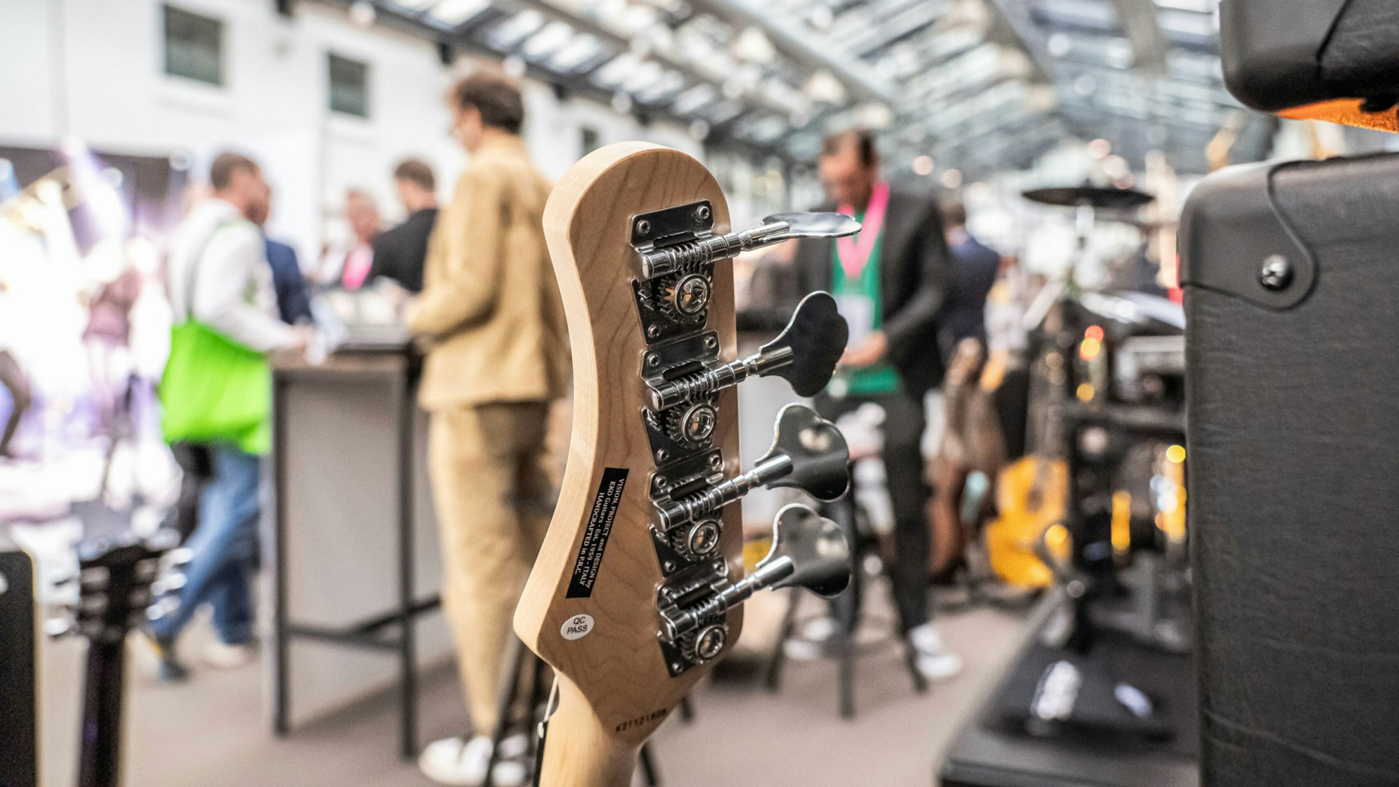 Teile von einer Gitarre sind im Vordergrund, im Hintergrund halten sich mehrere Personen in einer Halle auf.