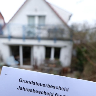 Ein Grundsteuerbescheid für 2024 wird vor einem Privatgrundstück hochgehalten.