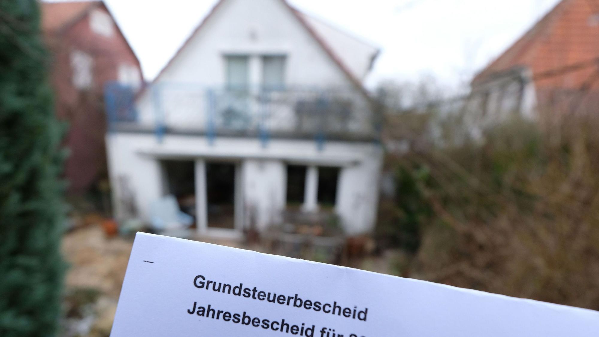Ein Grundsteuerbescheid für 2024 wird vor einem Privatgrundstück hochgehalten.