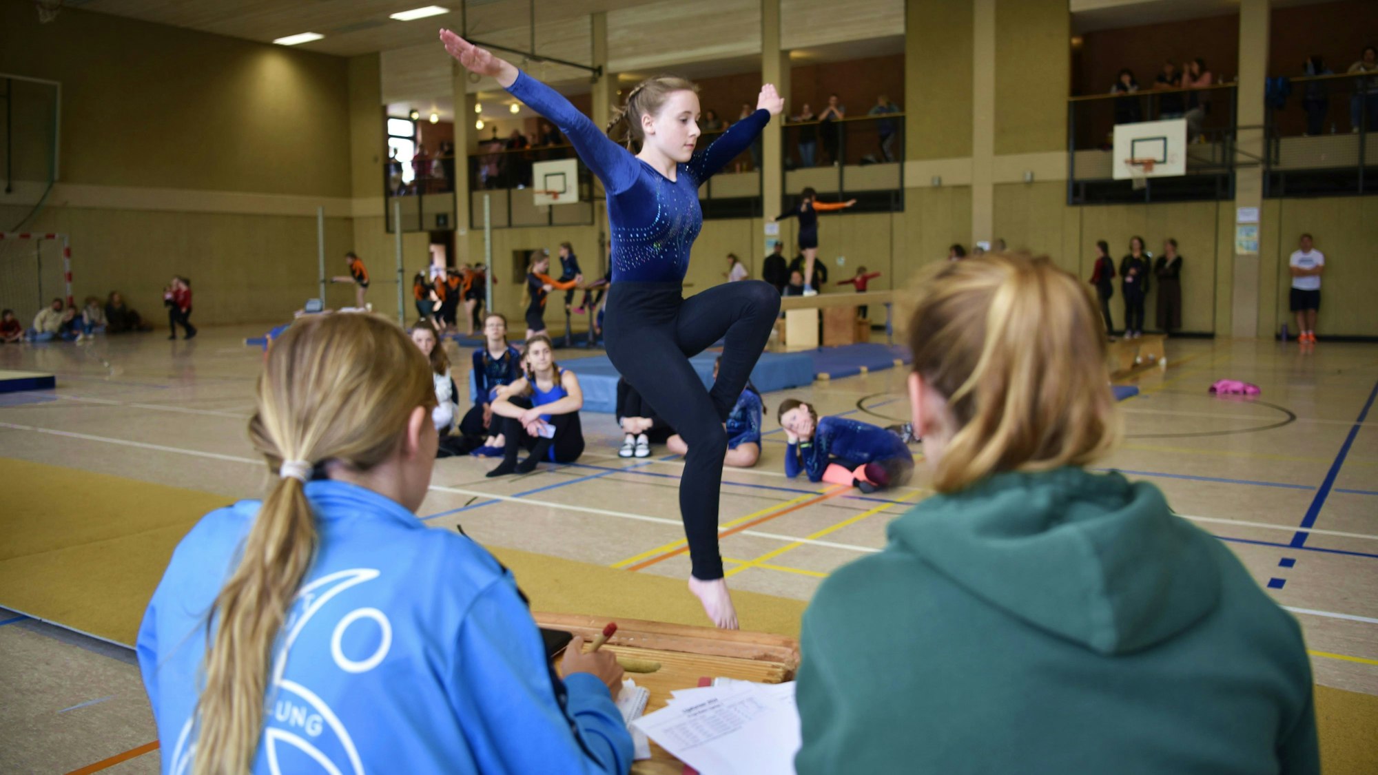 Eine Turnerin des Tus Schleiden zeigt beim Liga-Turnen ihre Bodenübung. Sie macht gerade einen Pferdchensprung und wird dabei von zwei Kampfrichterinnen sowie anderen Turnerinnen beobachtet.