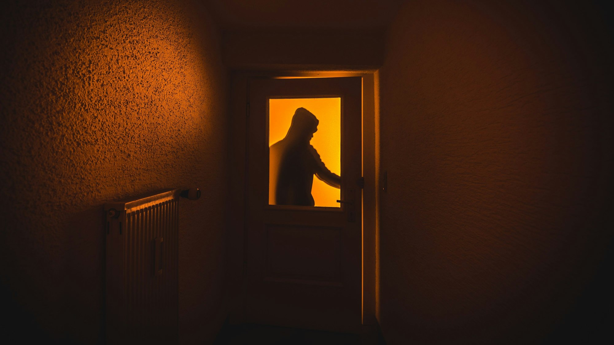 Durch eine Glasscheibe in einer Tür sieht man eine Person, die in eine dunkle Wohnung rein möchte.