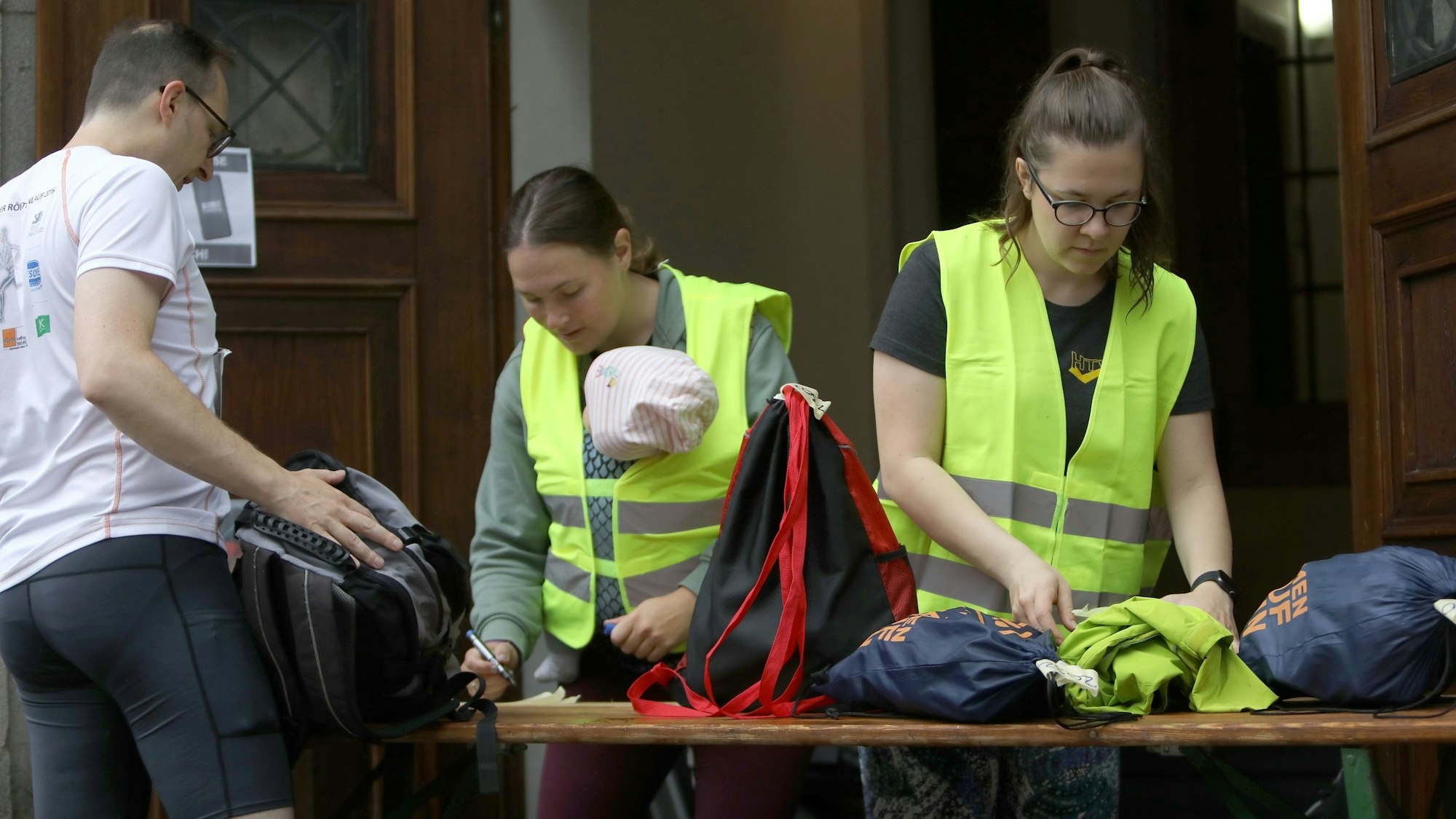 Zwei Frauen stehen an einem Tisch mit Rucksäcken, eine trägt ein Baby in ihrer Warnweste.