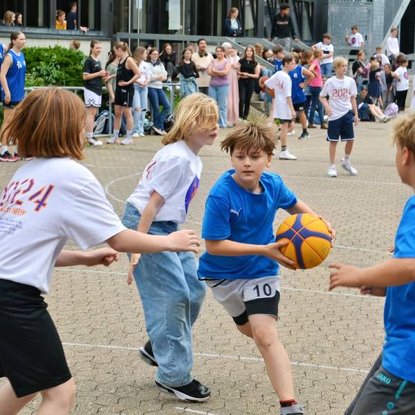 Mehrere Kinder spielen zusammen Basketball. Ein Junge mit blauem t-Shirt hat den Ball in den Händen.