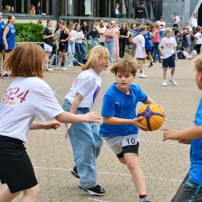 Mehrere Kinder spielen zusammen Basketball. Ein Junge mit blauem t-Shirt hat den Ball in den Händen.