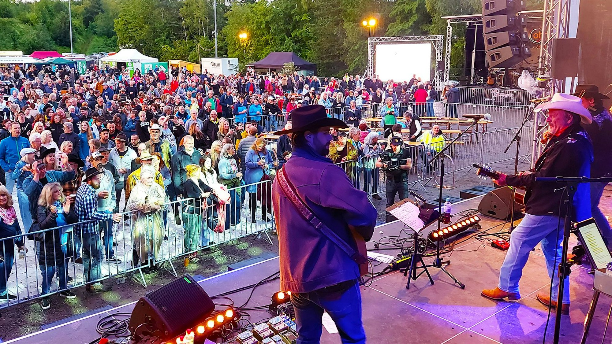 Männer in Cowboy-Hüten stehen auf einer Bühne und spielen Gitarre. Vor der Bühne haben sich zahlreiche Menschen versammelt.
