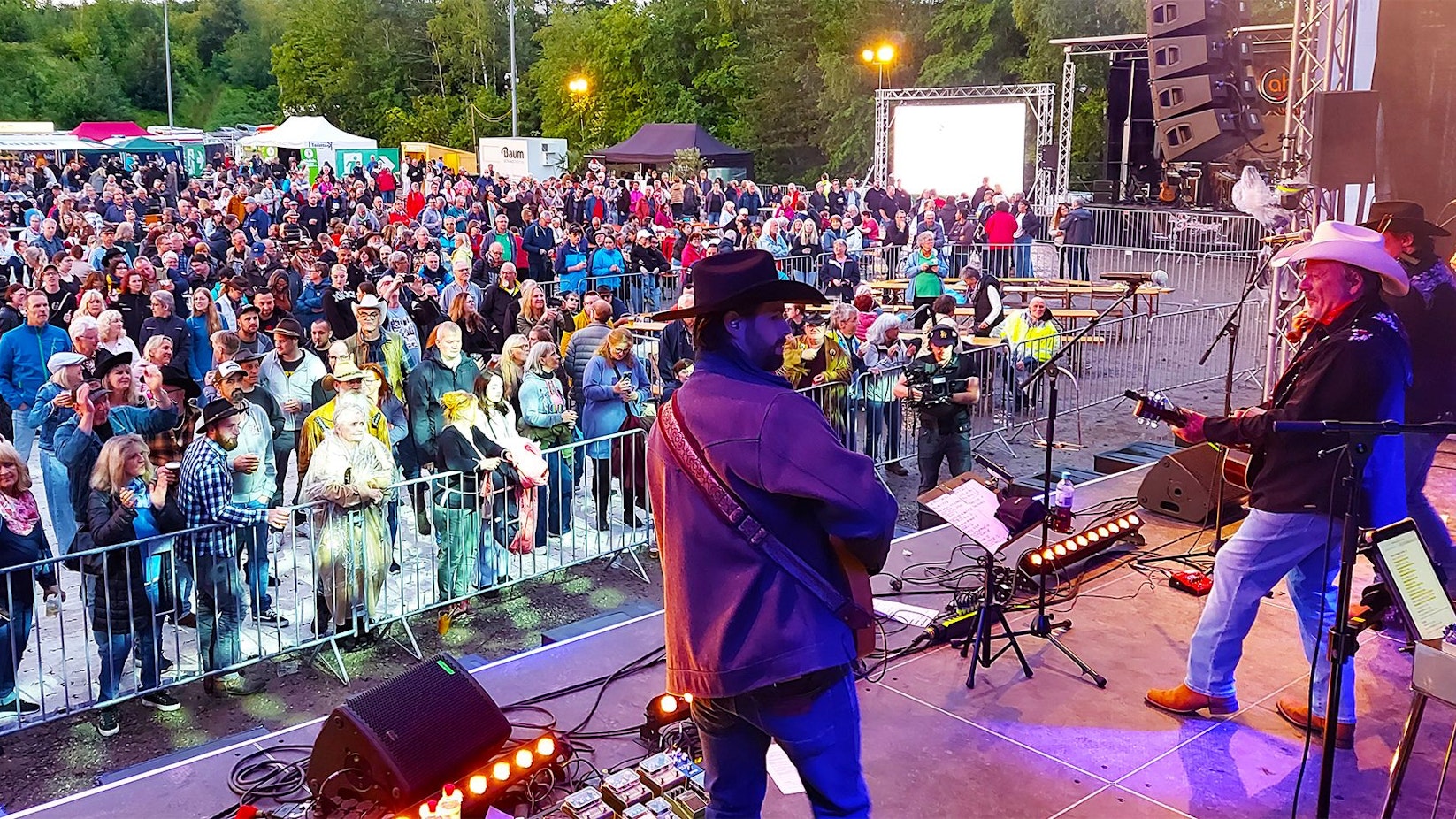 Männer in Cowboy-Hüten stehen auf einer Bühne und spielen Gitarre. Vor der Bühne haben sich zahlreiche Menschen versammelt.