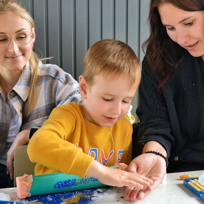 Ein Kind bastelt mit Glitzer, zwei Frauen helfen ihm.
