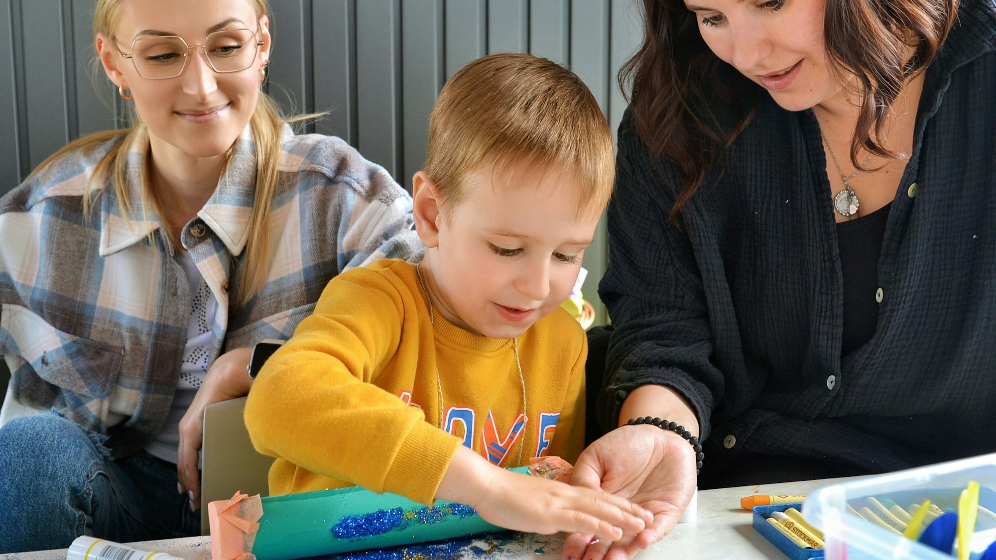 Ein Kind bastelt mit Glitzer, zwei Frauen helfen ihm.