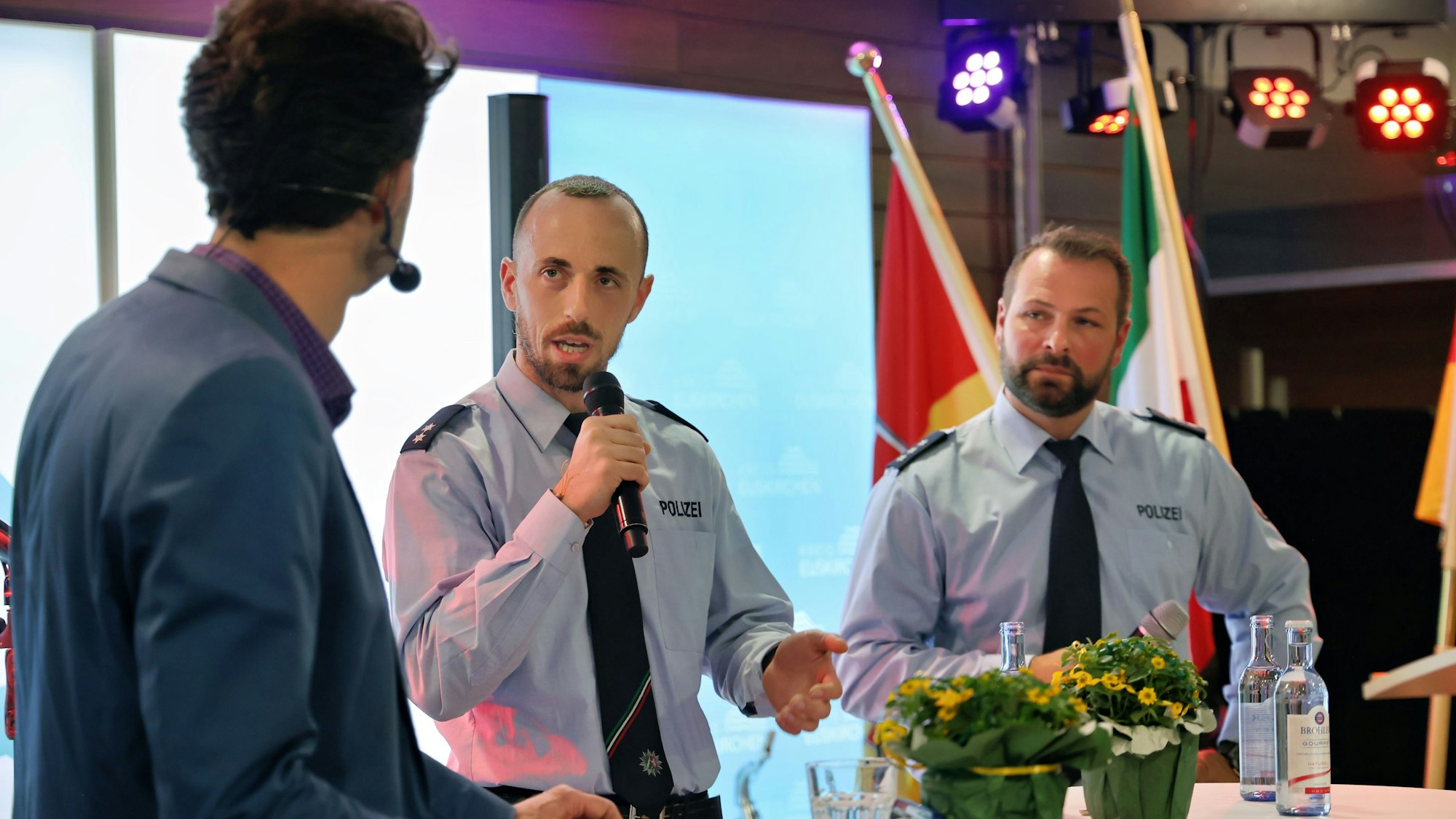 Zwei Polizisten in Uniform stehen an einem Stehtisch und sprechen mit einem Moderator.