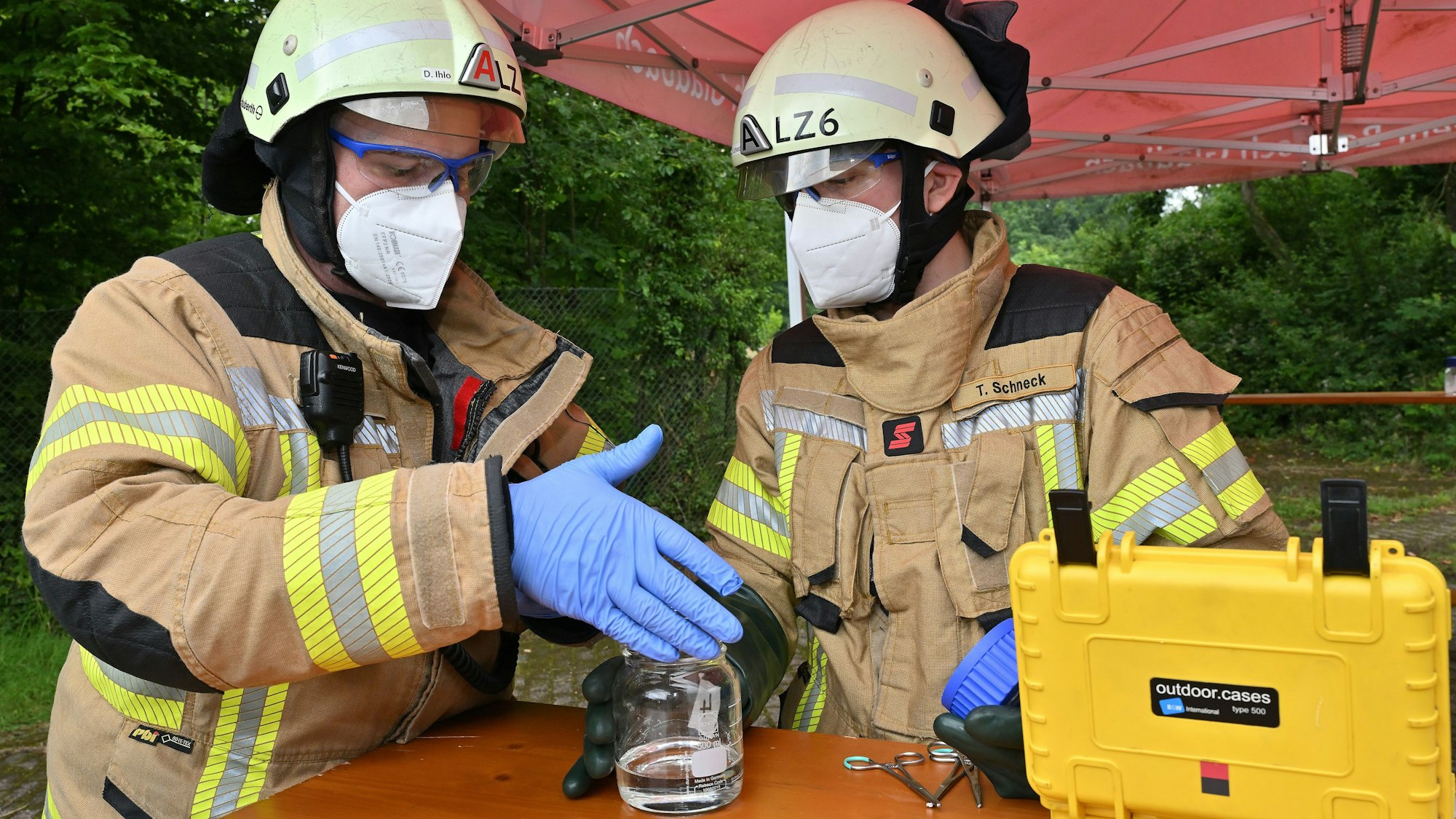 Feuerwehrleute mit Mund-Nase-Schutz und Schutzbrillen analysieren eine Flüssigkeit in einem Glas.