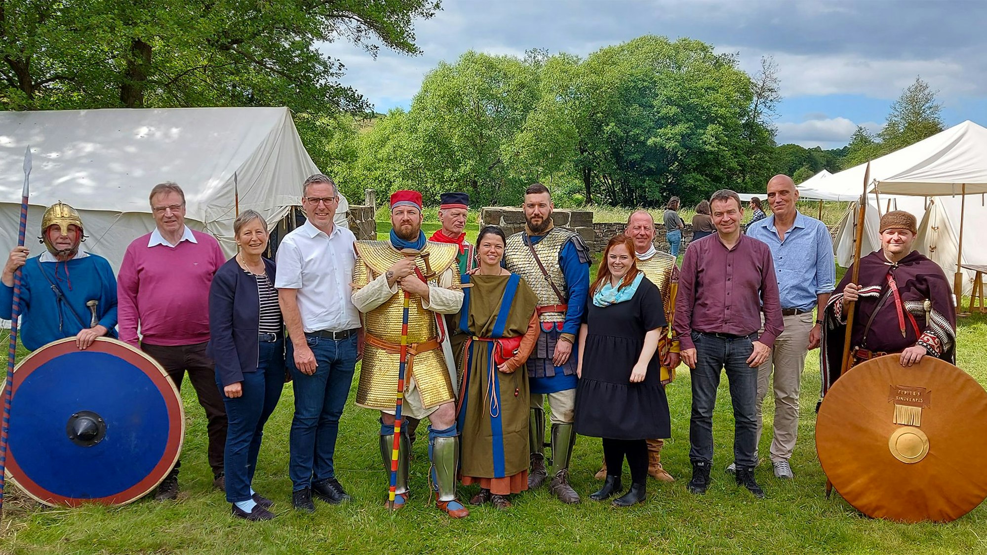 Eine Gruppe von Menschen steht zwischen weißen Zelten auf einer Wiese, einige tragen historische Kostüme.