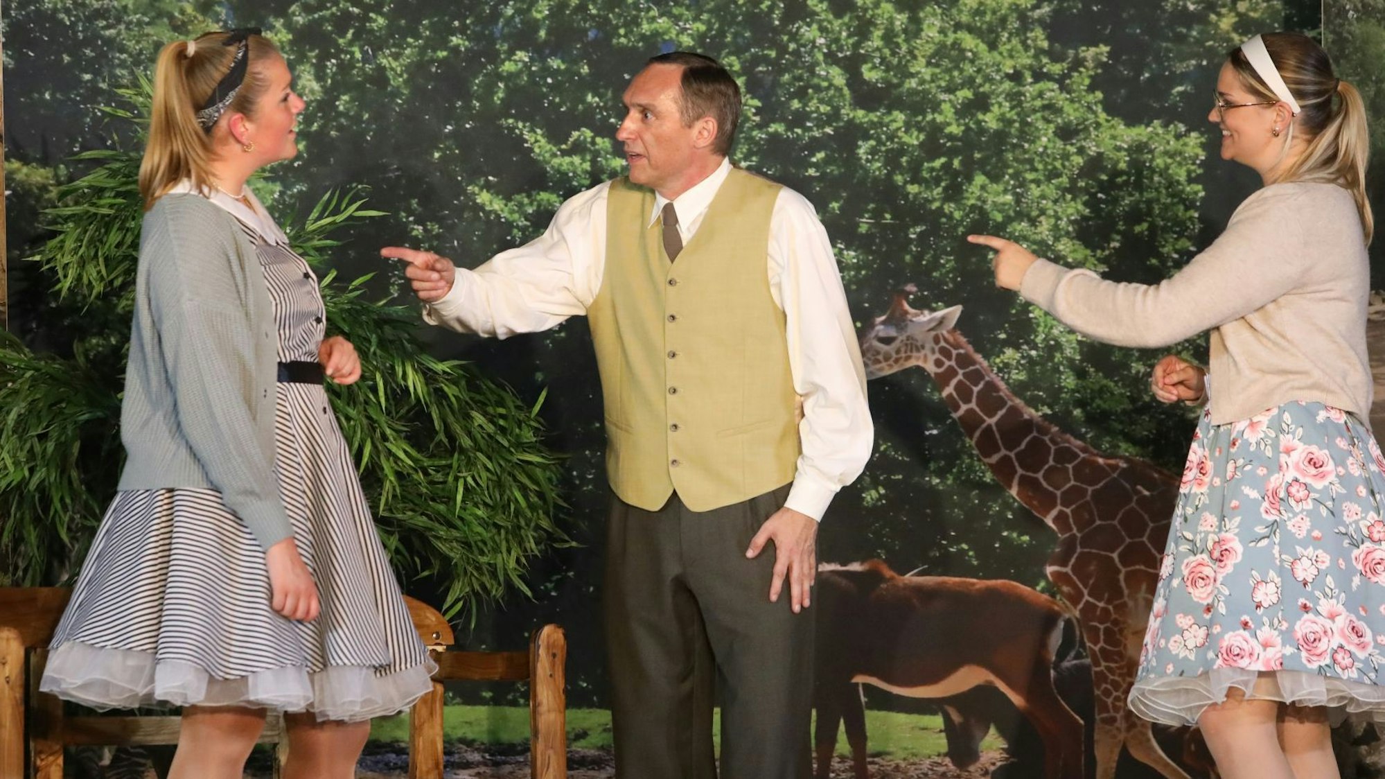 Zwei Frauen und ein Mann stehen auf einer Bühne, im Hintergrund ist einer Wand eine Giraffe zu sehen.