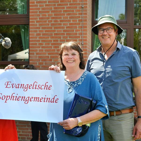 Zwei Frauen halten ein Schild mit der Aufschrift „Evangelische Sophiengemeinde“ fest, daneben steht ein Mann, der einen grünen Anglerhut trägt.