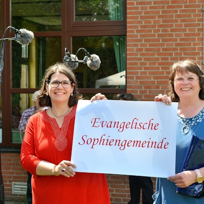 Zwei Frauen halten ein Schild mit der Aufschrift „Evangelische Sophiengemeinde“ fest, daneben steht ein Mann, der einen grünen Anglerhut trägt.