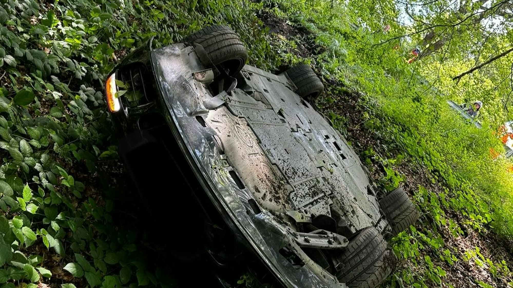 Ein Porsche Cabrio liegt auf dem Dach im Wald, ein Frontscheinwerfer leuchtet.