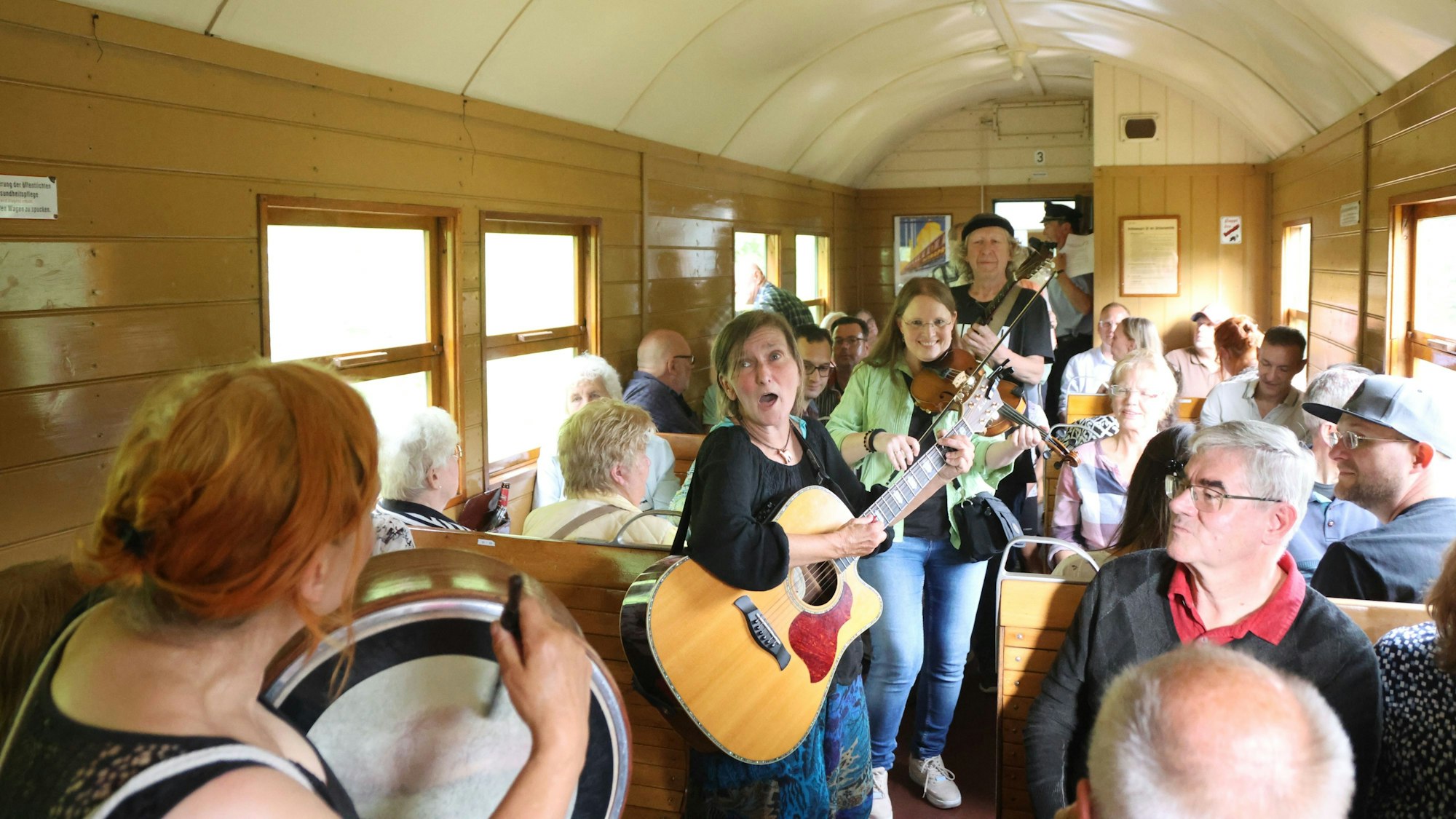 Das Innere eines historischen Eisenbahnwaggons zeigt viele Menschen und Musiker mit Gitarre und weiteren Instrumenten.