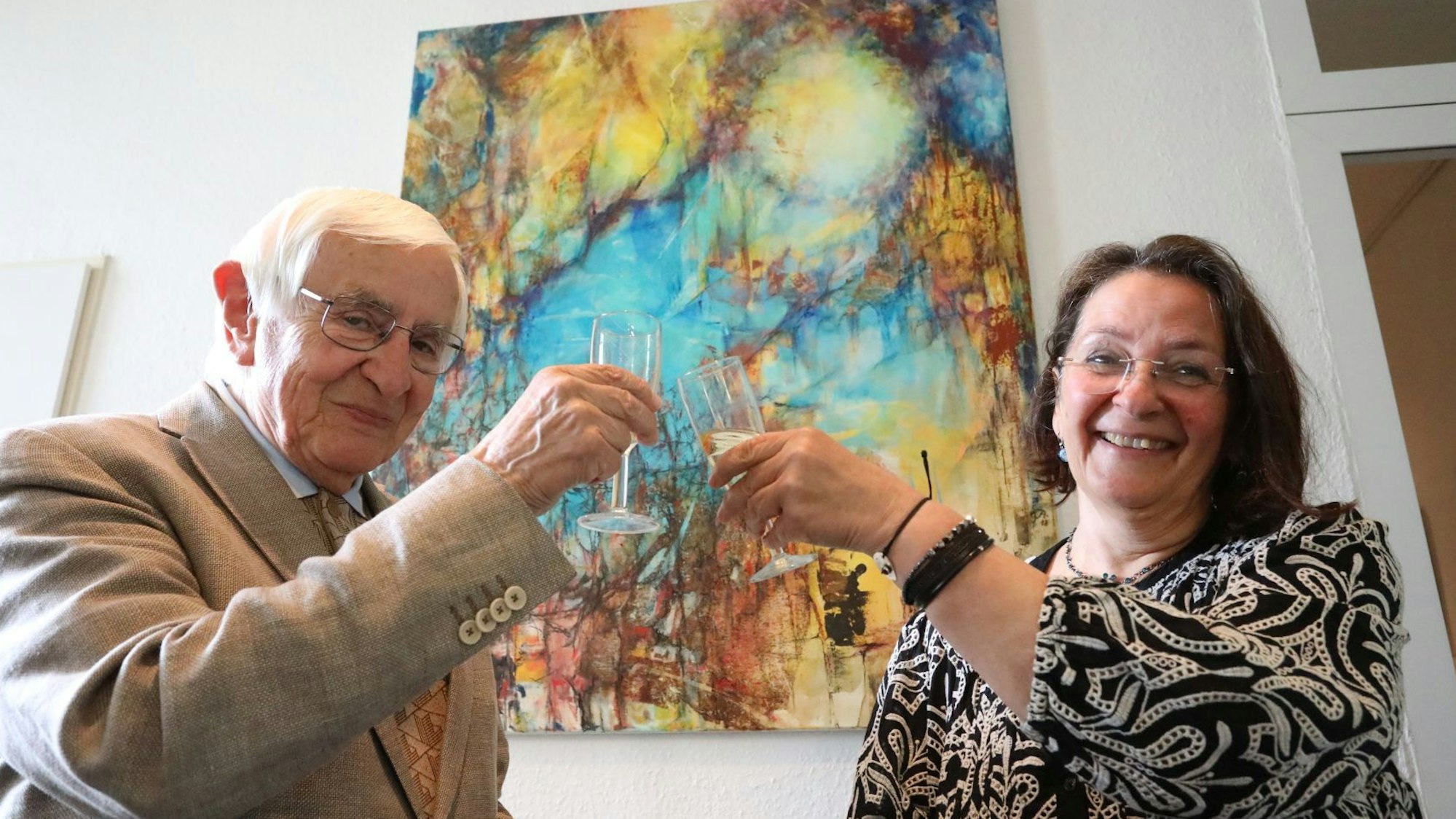 Ein Mann und eine Frau stoßen vor einem Kunstwerk mit einem Glas Sekt an.