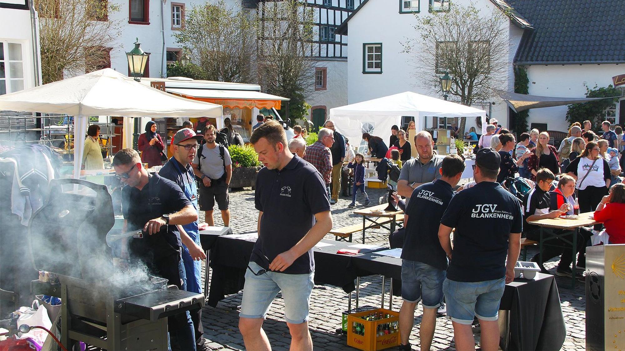 Einige junge Männer stehen im historischen Ortskern von Blankenheim an einem Grill. Im Hintergrund sind zahlreiche Besucher und weitere Stände zu sehen.