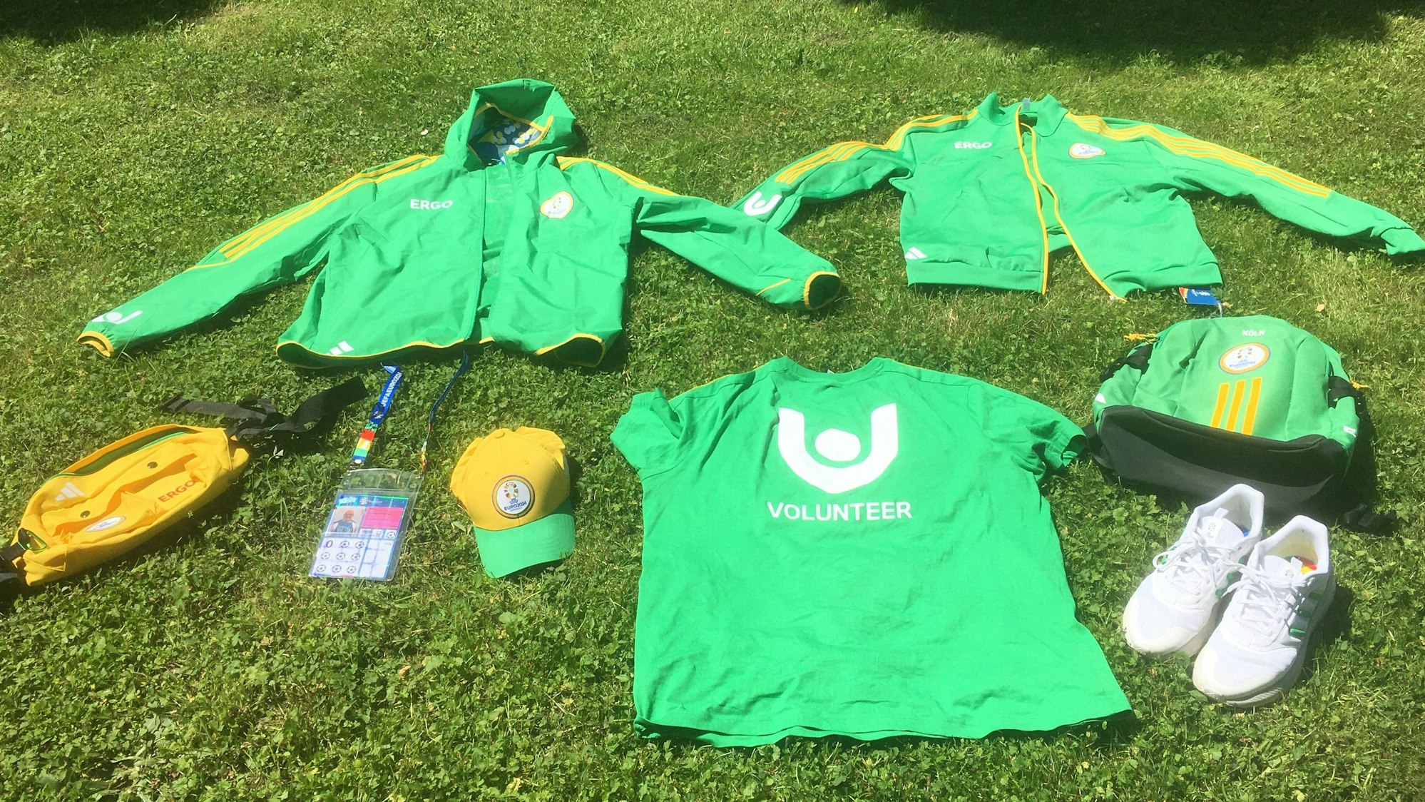 Auf einen Rasen sind Kleidungsstücke und Ausstattung eines EM-Volunteers ausgebreitet: grünes Shirt, grüne Jacken, weiße Schuhe, gelbes Käppi, grüner Rucksack, Ausweis und gelbe Bauchtasche.