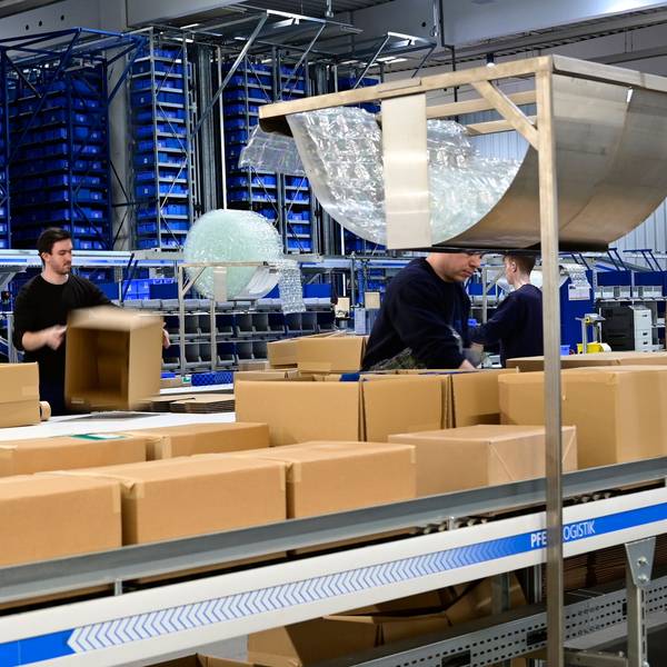 Das Foto zeigt eine Halle der Firma Rüggeberg mit vielen Paketen und einigen Mitarbeitern