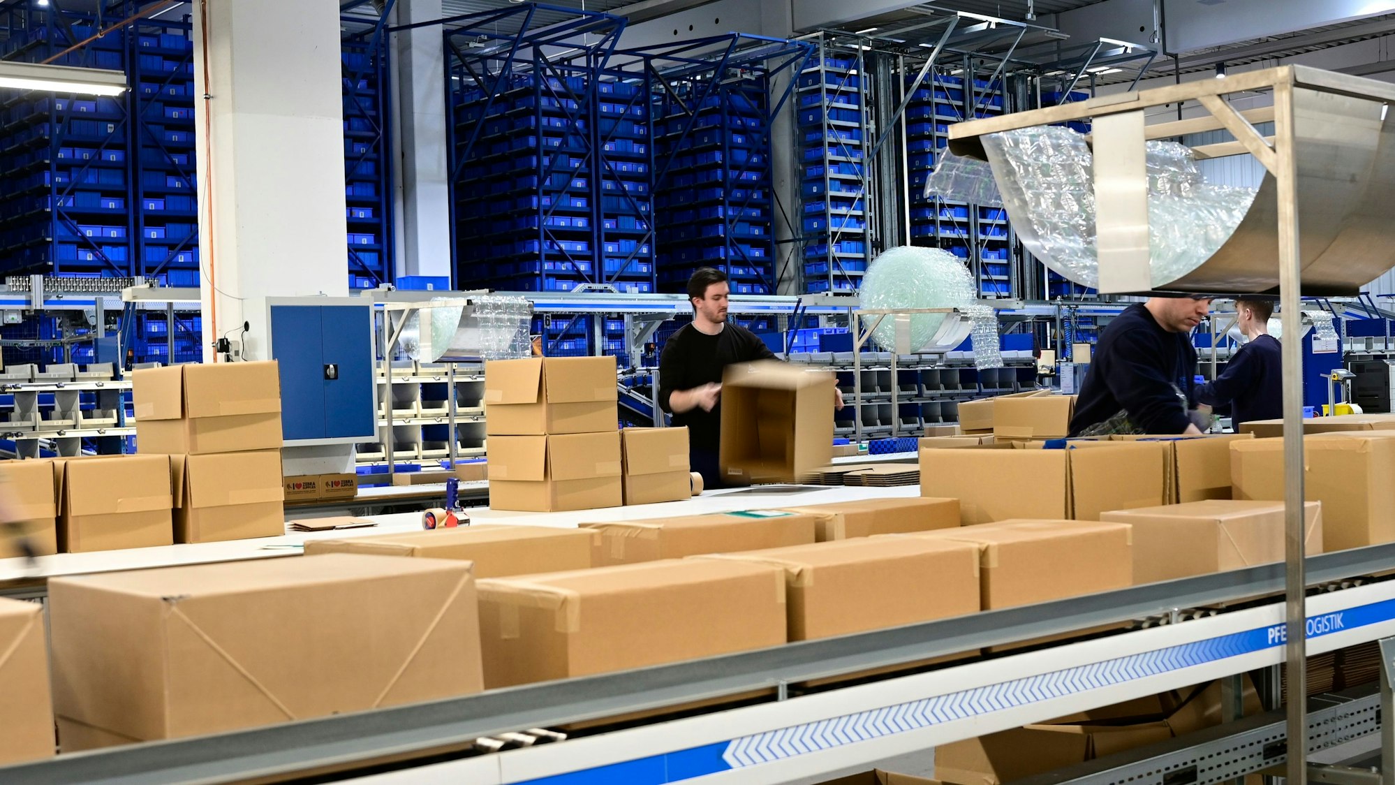 Das Foto zeigt eine Halle der Firma Rüggeberg mit vielen Paketen und einigen Mitarbeitern