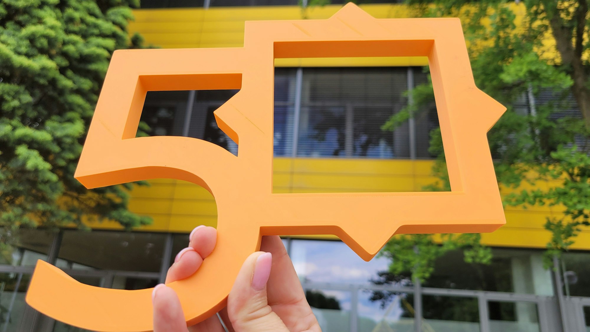 Eine Hand hält die Zahl 50 hoch. Sie wurde mit einem 3D-Drucker angefertigt. Im Hintergrund ist die markante gelbe Fassade des Schulgebäudes zu sehen.
