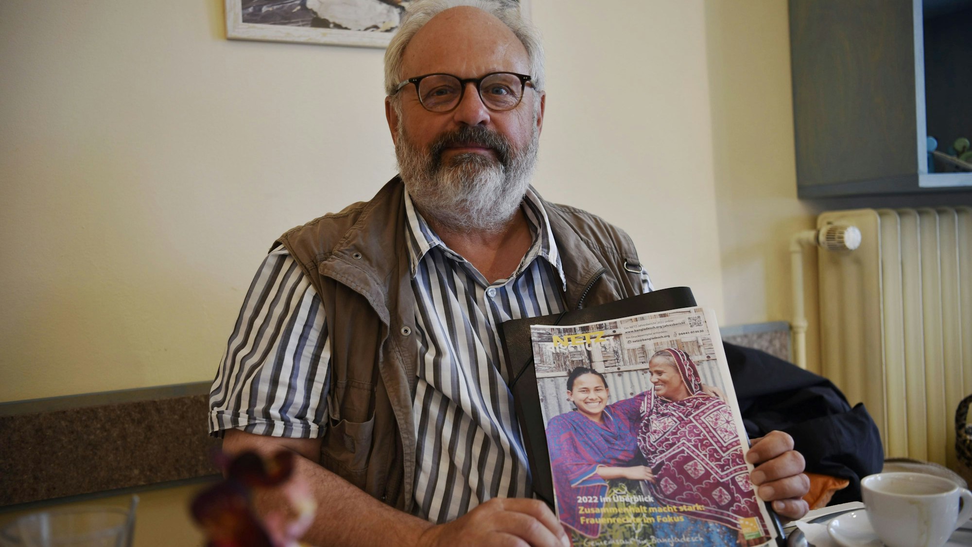 Joachim Berg sitzt im Fair Café in Zülpich und hält einen Flyer der Hilfsorganisation Netz Bangladesch in die Kamera.