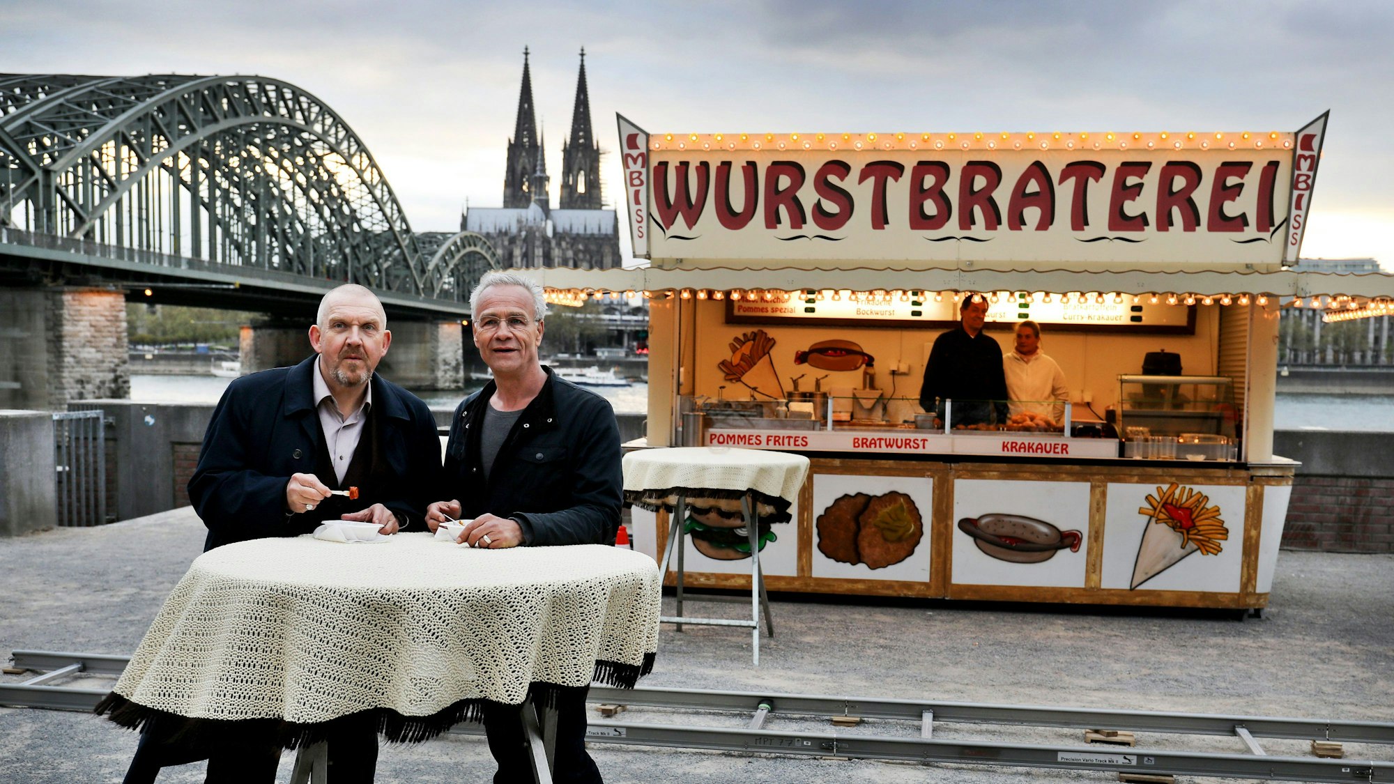Die Tatort-Schauspieler Dietmar Bär und Klaus J. Behrendt (r.) stehen in Köln vor Dreharbeiten an der Wurstbraterei vor dem Dom.