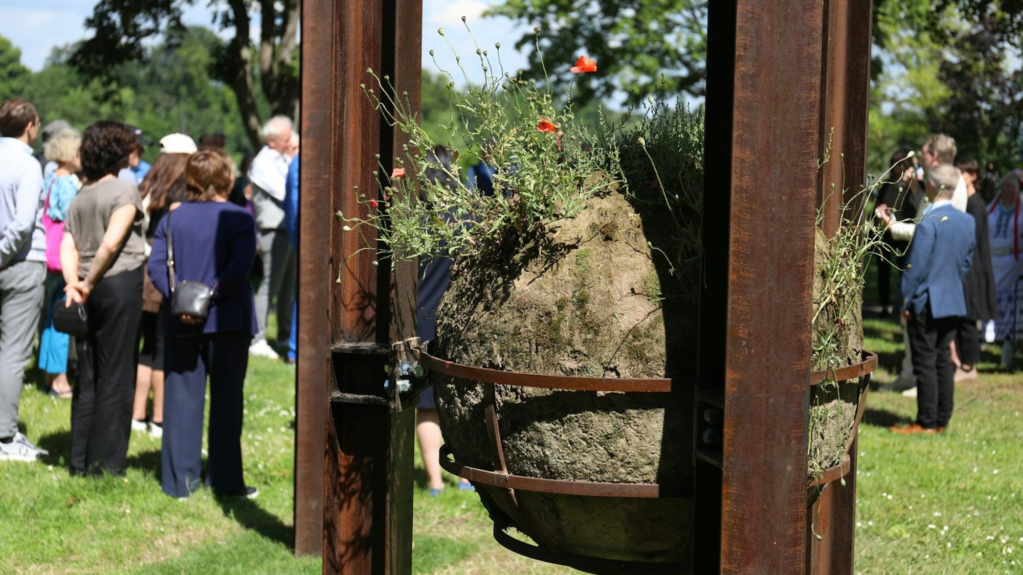 In einem Metallkorb, der an Stahlstelen befestigt ist, liegt ein großer Lehmball. Aus ihm sprießen Mohnblumen.