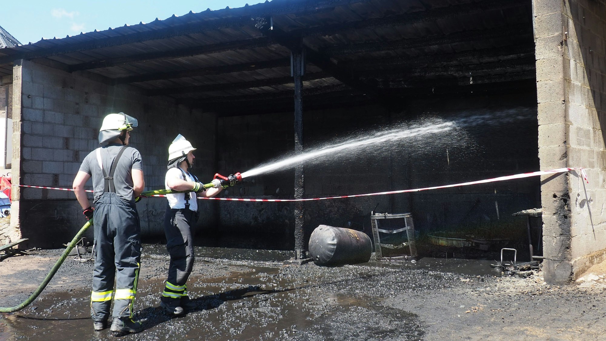 Ein Feuerwehrmann spritzt Wasser in die ausgebrannte Halle, ein anderen hält den Schlauch.